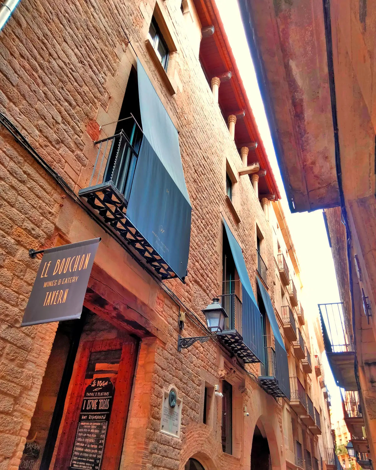 Stone facade of Hotel Mercer on Carrer dels Lledó: dark awnings, iron balconies and the Le Bouchon tavern sign