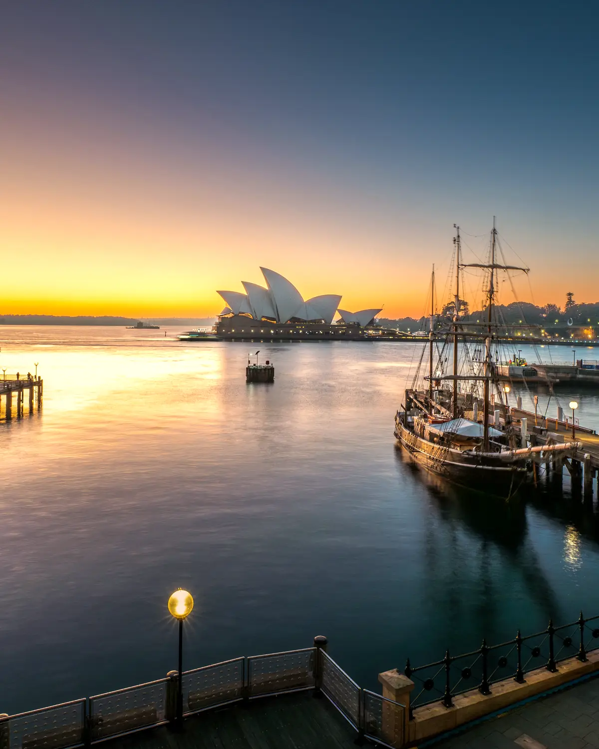 Sydney Opera House at sunrise from a harbour terrace, with a lamp and a sailing ship in the cove