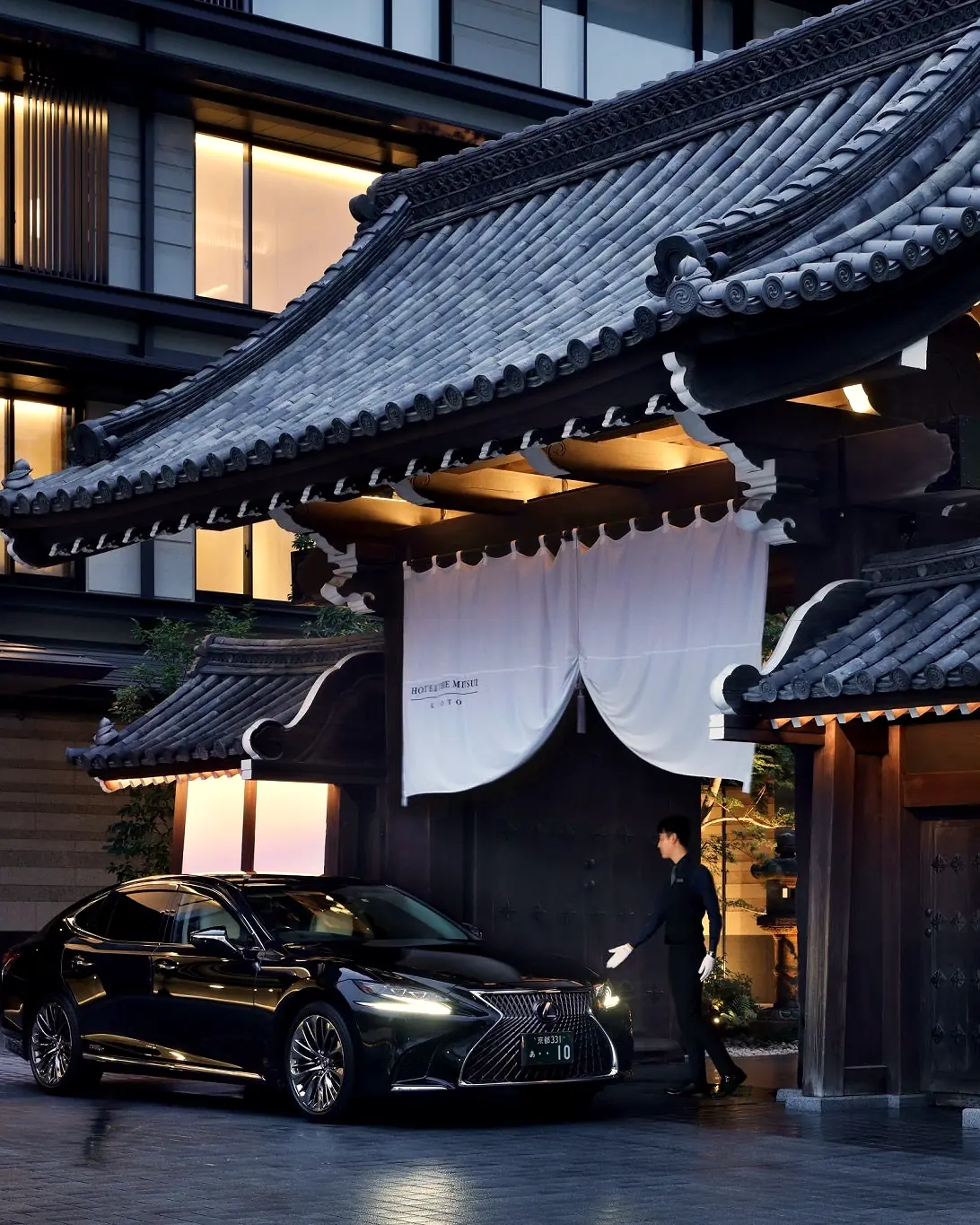 The tiled-roof entrance gate with a white noren curtain at dusk, a car and doorman in front and the hotel building behind
